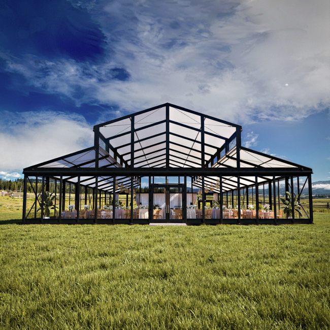 Glass-walled pavilion with a peaked roof sits on a grassy field under a blue sky, set for an outdoor event with tables and chairs inside.