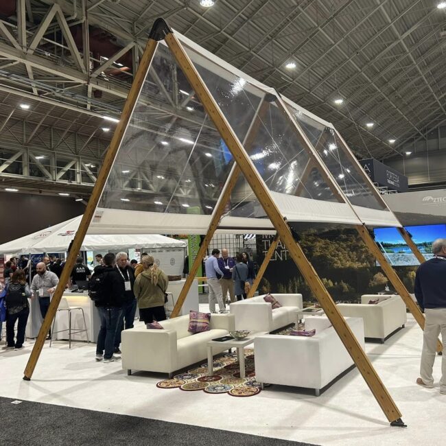 Wooden triangular A-frame canopy with clear panels over a white lounge seating area at a trade show event.