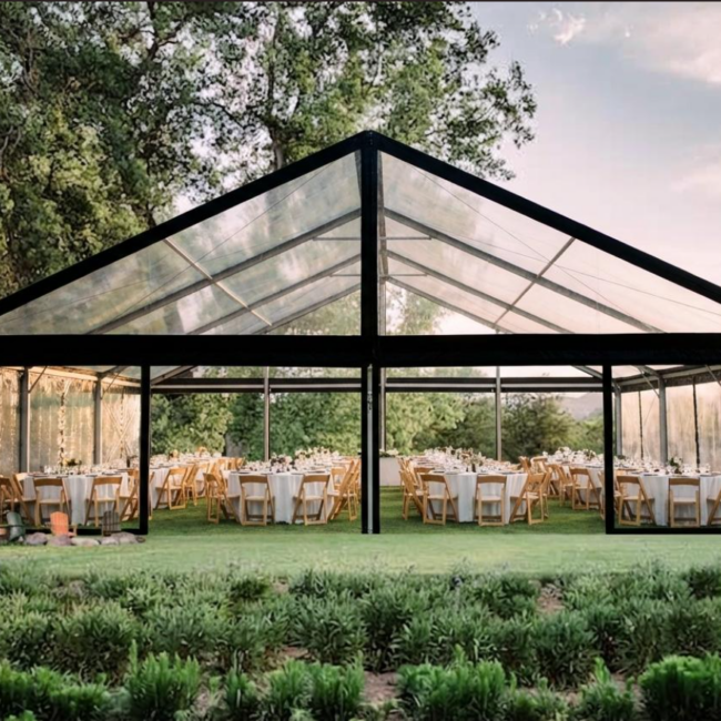 Outdoor glass-walled event tent set up with round dining tables and chairs on a green lawn, surrounded by trees at dusk.