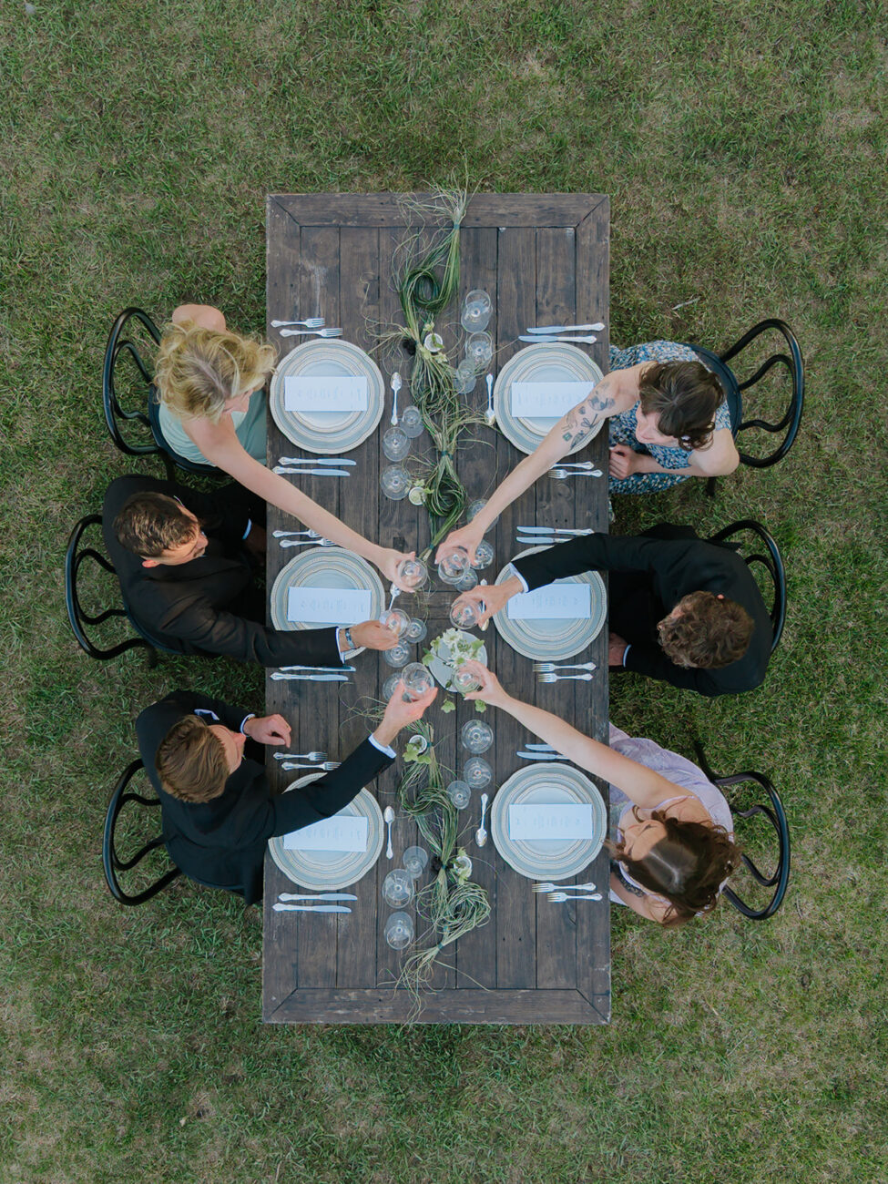 Overhead shot of eight people around a rustic wooden table on grass, toasting with wine glasses during an outdoor dinner party.