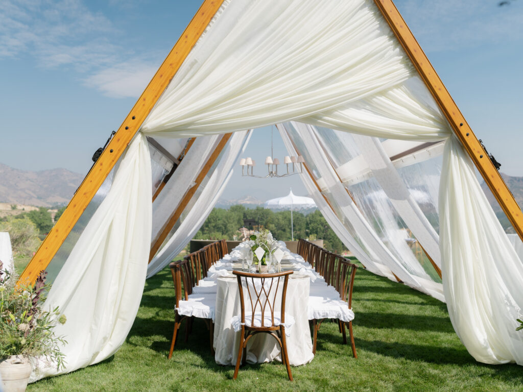 Outdoor wedding reception under wooden A-frame arches with white drapes, a long banquet table, and wooden chairs on a green lawn.