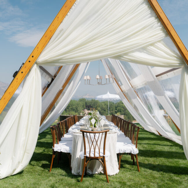 Outdoor wedding setup under white draped canopy with wooden chairs and a long, linen-covered table on a green lawn, mountains in the background.
